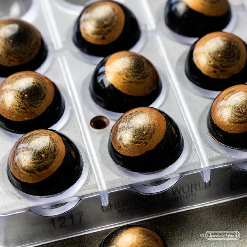 Glossy dark chocolate dome-shaped bonbons decorated with brushed gold cocoa butter, set in a clear Chocolate World mould.