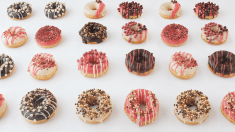 Assorted mini doughnuts decorated with colourful chocolate drizzles and textured Mona Lisa sprinkles in pink, white, gold and dark chocolate tones, arranged in a repeating pattern on a white background.