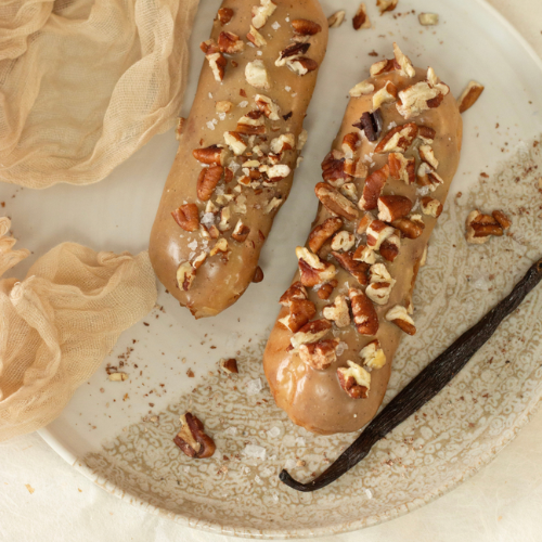 Two pecan vanilla éclairs topped with caramel-coloured Callebaut Gold glaze and chopped pecans, served alongside a vanilla pod.