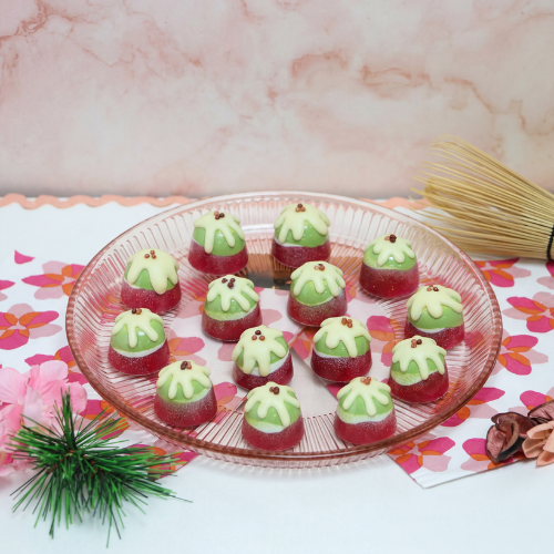 Festive matcha and cherry bonbons decorated to resemble miniature Christmas puddings, featuring glossy green matcha tops, cherry-red shells and white chocolate detailing. Arranged on a glass plate with seasonal styling, these chocolates combine nostalgic festive inspiration with contemporary Asian flavours.