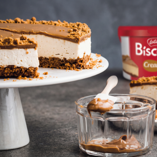 Slice of creamy Biscoff cheesecake topped with Lotus Biscoff spread and biscuit crumb on a white cake stand, alongside a glass bowl of smooth Biscoff spread and a Lotus Biscoff tub in the background.