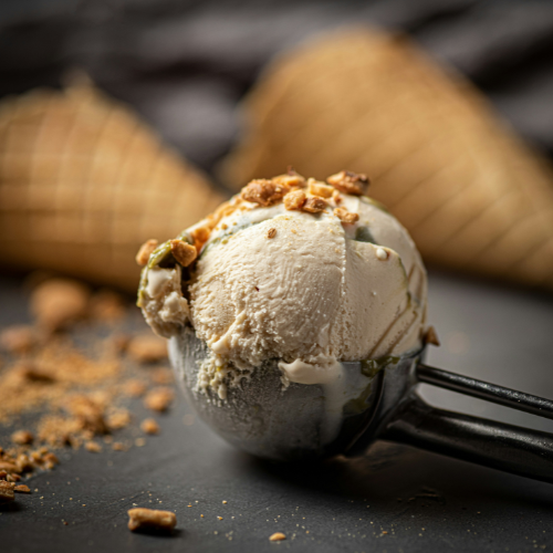Close-up of a scoop of creamy vanilla ice cream in a metal ice cream scoop, topped with crunchy caramelised biscuit crumbs. Two waffle cones are softly blurred in the background, with scattered crumbs across a dark surface.