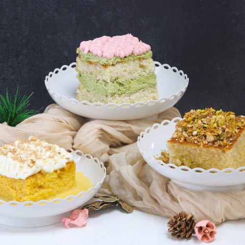 Three festive vanilla sponge cakes presented with different flavour profiles: matcha and cherry blossom, coconut and mango tres leches, and baklava-inspired orange honey and pistachio. The cakes are displayed on decorative stands with seasonal styling and festive decorations.