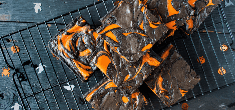 Halloween brownies topped with swirls of vivid orange chocolate and black chocolate, stacked on a cooling rack with ghost, pumpkin and spider decorations scattered around.