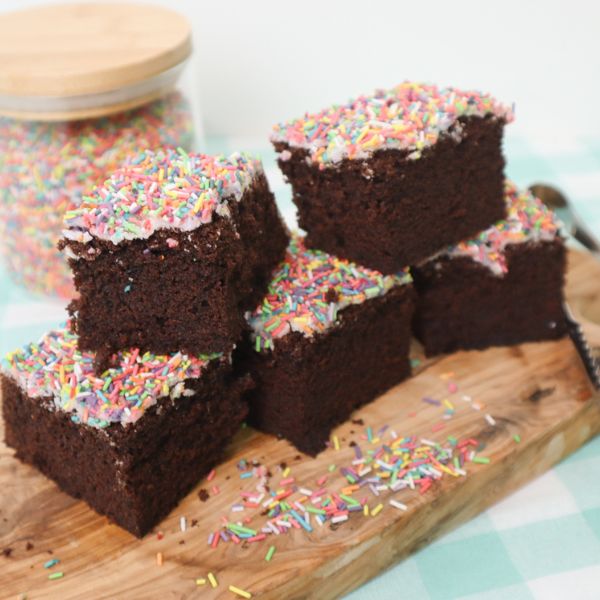 Chocolate cake squares topped with icing and colourful sprinkles stacked on a wooden board