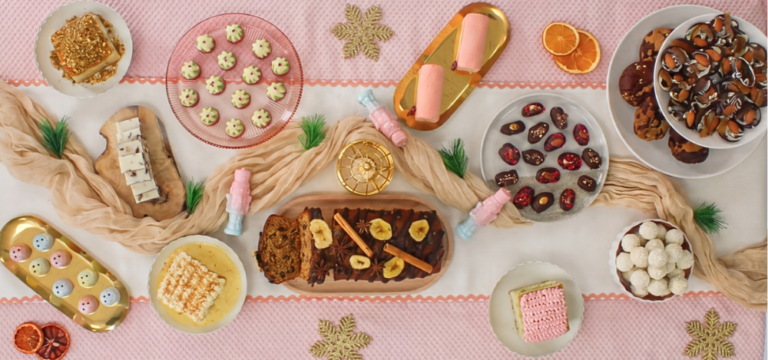 Overhead view of a festive Christmas dessert table featuring a variety of chocolates, cakes, confectionery and seasonal treats arranged amongst decorative Christmas styling, gold accents and winter-themed decorations.