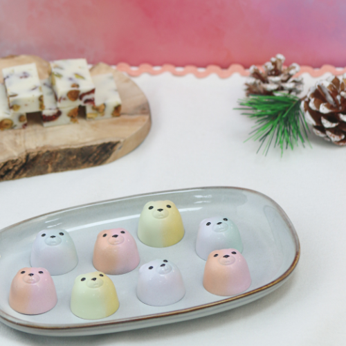 Pastel-coloured bear-shaped chocolates decorated with Roxy & Rich coloured cocoa butters in soft pink, green and lilac tones. The festive bonbons are arranged on a serving plate with seasonal decorations and feature a miso caramel crunch filling.