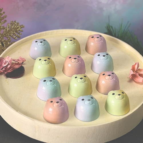 Pastel-coloured bear-shaped chocolates arranged on a wooden tray, featuring glossy finishes in pink, blue and green, surrounded by festive decorations and dried flowers.