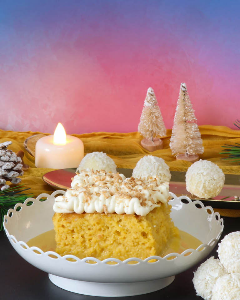 A festive display featuring a slice of coconut and mango tres leches cake topped with cream and toasted coconut, alongside coconut-coated mango truffles. The desserts are styled with Christmas decorations, candles and miniature trees against a pink and blue background.
