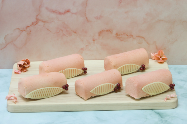 Five mini pink yule log desserts coated in a velvety finish, decorated with white chocolate leaf details and red berries, displayed on a wooden board against a soft pink marble backdrop.