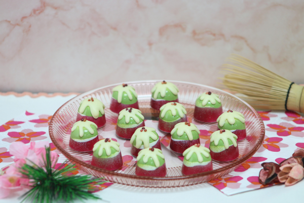 Matcha and cherry blossom cheesecake bonbons decorated in festive green and pink tones, arranged on a glass plate with cherry blossom-inspired styling and seasonal decorations. The bonbons feature glossy matcha tops, cherry-flavoured shells and delicate white chocolate detailing.
