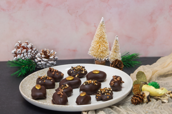 Chocolate-coated dates filled with banana-flavoured ganache and decorated with Mona Lisa chocolate sprinkles and cocoa nibs. The festive treats are arranged on a plate alongside Christmas trees, pinecones and seasonal decorations.