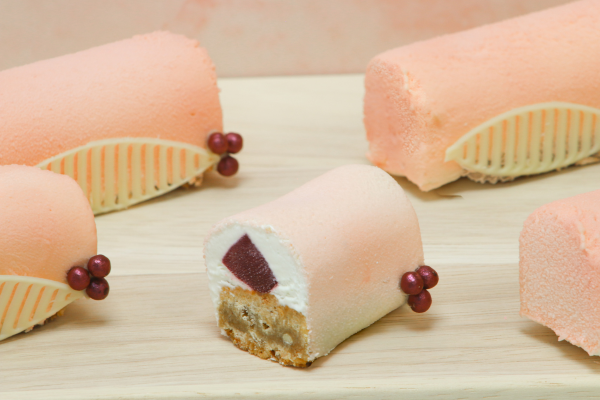 Five mini pink yule log desserts coated in a velvety finish, decorated with white chocolate leaf details and red berries, displayed on a wooden board against a soft pink marble backdrop.