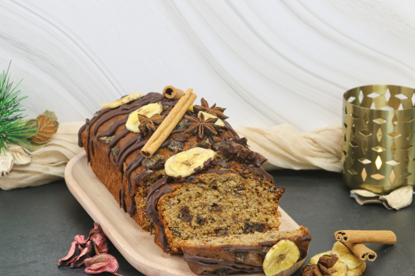 A festive vegan loaf cake made with banana, dates and dark chocolate, decorated with banana chips, cinnamon sticks and star anise. The loaf is presented on a wooden serving board with seasonal Christmas styling and winter decorations.