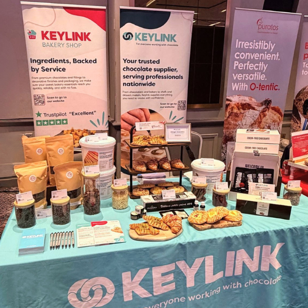 Keylink exhibition stand at a Scottish Bakers industry event, displaying bakery ingredients, chocolate products, inclusions, fillings and sample pastries. Branded Keylink banners promote bakery and chocolate solutions, while product samples and finished baked goods are showcased on a turquoise Keylink tablecloth.