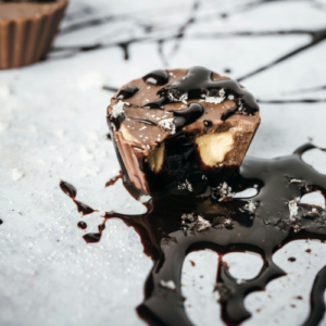 Chocolate cup drizzled with dark chocolate sauce and finished with flakes of sea salt. One chocolate cup has been cut open to reveal a smooth, creamy filling, creating an indulgent sweet and salty confectionery treat.