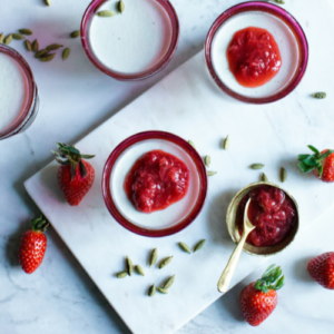Individual dessert pots filled with a smooth, creamy dessert and topped with strawberry compote. Fresh strawberries and scattered cardamom pods surround the desserts, highlighting a flavour combination of sweet strawberry and aromatic cardamom in an elegant plated presentation.
