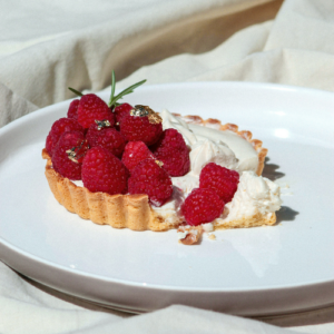 Individual tart filled with smooth white chocolate cream and topped with fresh raspberries, finished with gold leaf and a sprig of rosemary. The dessert showcases a premium flavour combination of raspberry, white chocolate and cardamom, presented on a white plate.